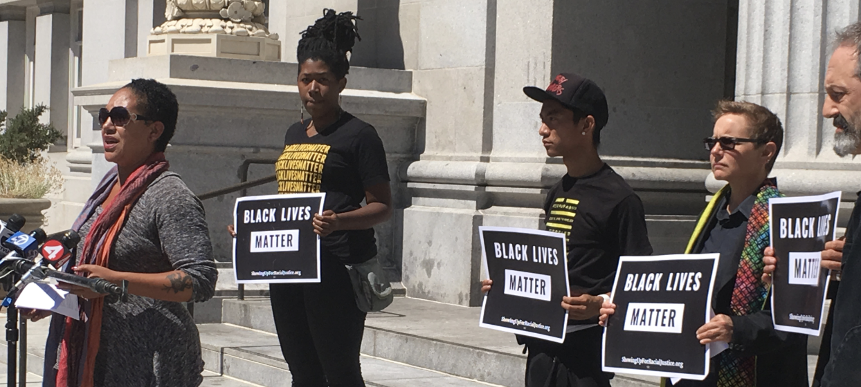 A multi-racial group hold Black Lives Matter signs behind Cat Brooks who is speaking at a press conference.Picture