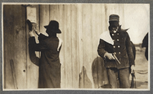 Old black and white photos of two men wearing hats, dark face coverings and white shoulder sashes. One man is putting a notice on the side of a barn, while the other man holds a gun in one hand and the reins of two horses in the other.