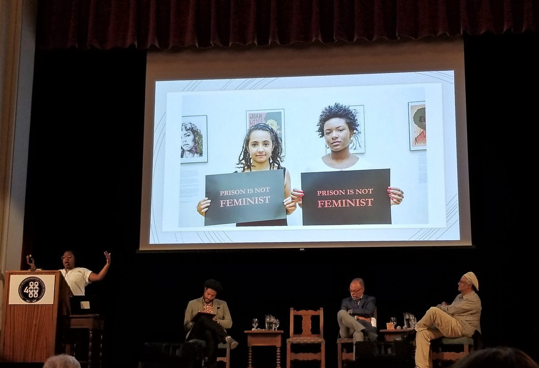 The panel on ‘Power and Resistance’ with C. Carruthers speaking at the podium and seated (left to right) J.S. Lewis (moderator), C. Henry and W. Martin, beneath a slide showing two young Black women carrying signs that say “Prison is not feminist.” (Photo by M. Luckey)Picture