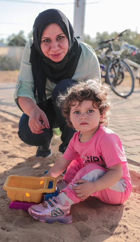 PictureWafaa and her niece Maryam at a MECA playground in Khuza’a, Gaza, in 2021.  The photo shows a woman squatting by a child with a toy on a sandy playground with a bicycle in the background. 